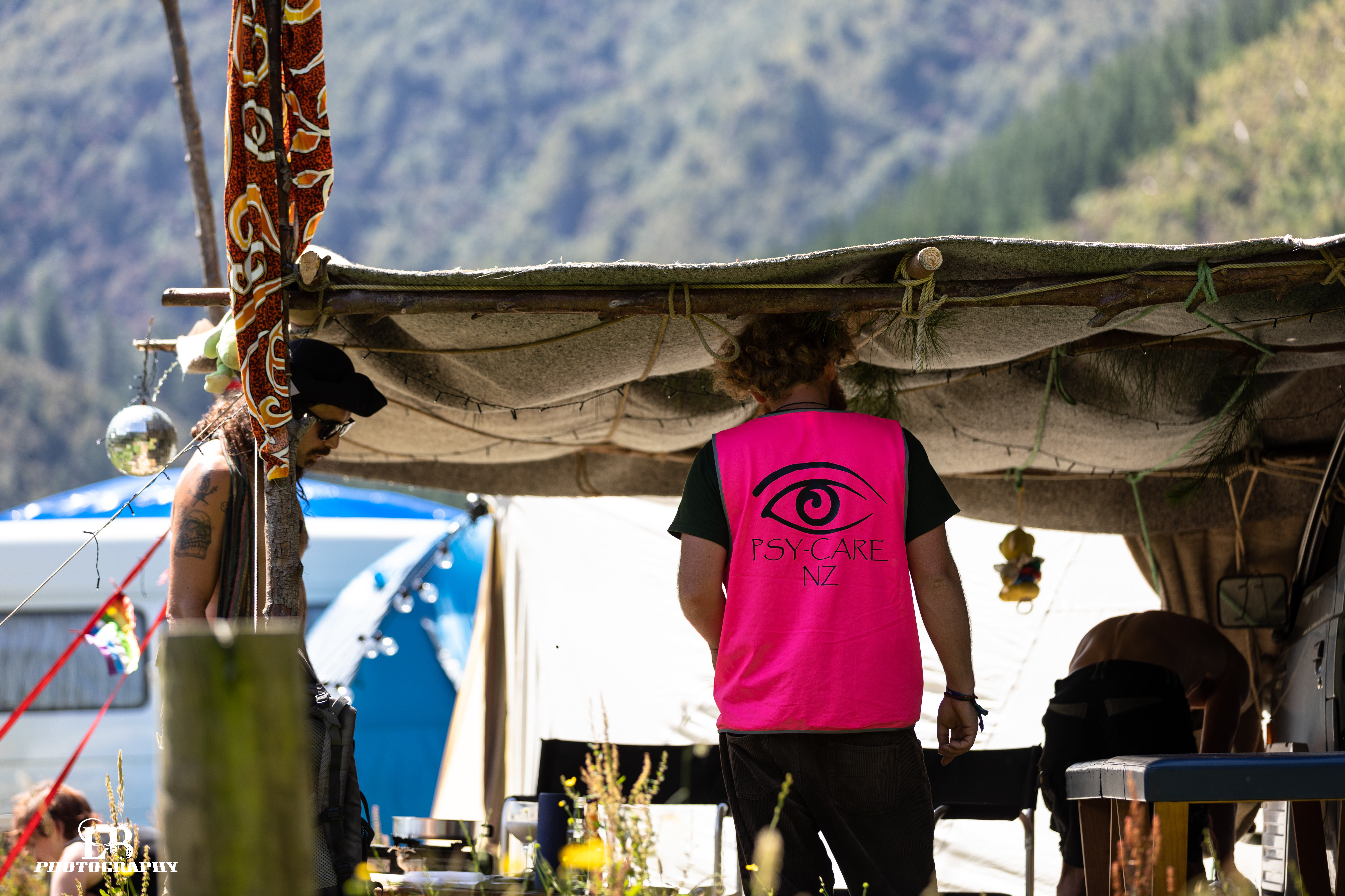 A person with their back to the camera, the Psy-Care NZ logo standing out on their pink hi vis vest. They are under an awning on a sunny day, and there are steep hills in the background.