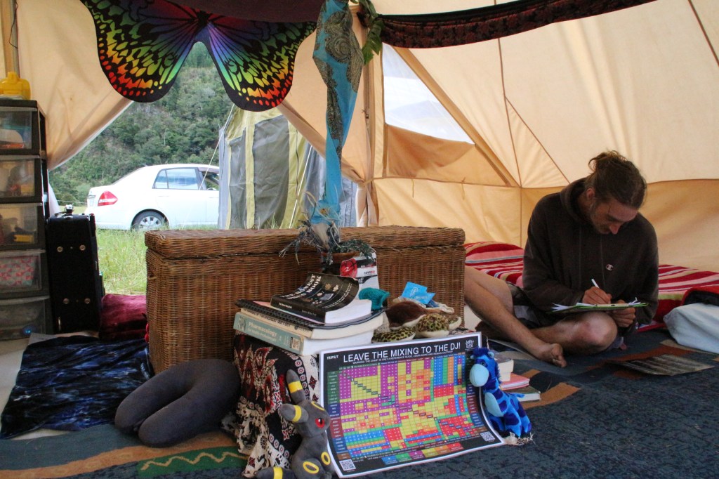 Inside one of the Psy-Care tents. A reference chart of drug interactions and several books are stacked on a small table. The tent is decorated with colourful fabric and several soft toys. A volunteer is sitting cross-legged filling out a clipboard.